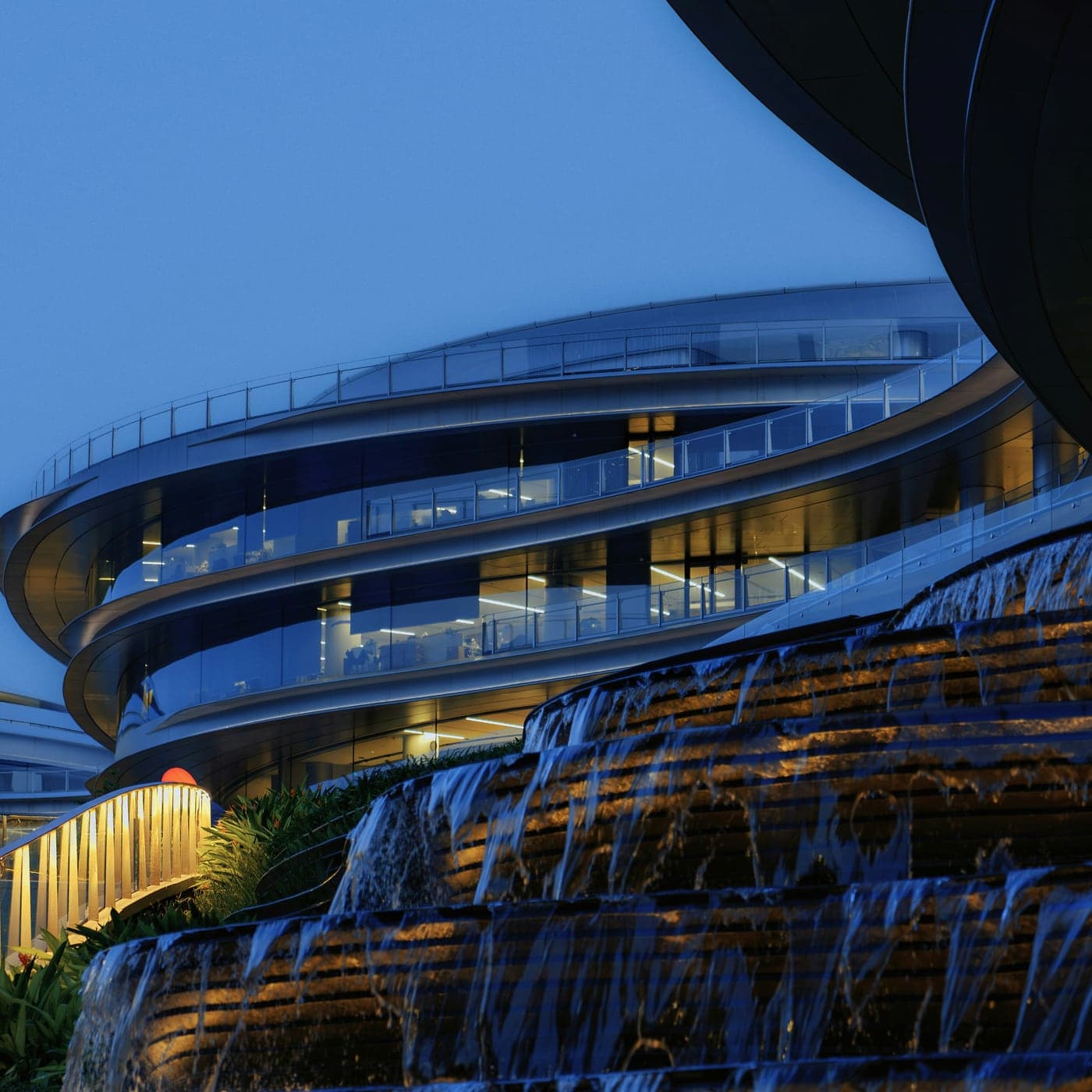 Curved glass office building at blue hour, occupied floors lit from within above a terraced water feature
